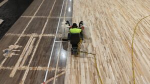 Worker operating a floor sander on a wooden gym floor, with yellow power cord trailing behind.