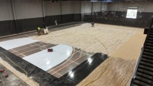 Indoor basketball court undergoing floor refinishing; workers sand and reseal the wooden floor with cables and equipment visible all around.