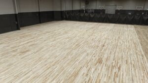 Indoor basketball court in a gym, with a light wooden floor and several hoops along the far wall. Black padded barriers line the perimeter.