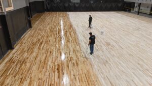 Two workers stand on a large varnished wood gym floor, one mopping and the other with a bucket nearby as finish is applied.