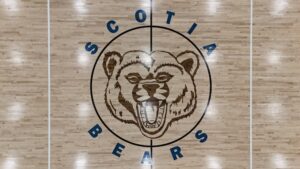 Wood gym floor with a large circular logo featuring a roaring bear head at center and blue letters arranged around the circle forming the team name.