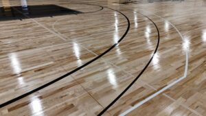 Empty indoor basketball court with a polished wooden floor and black and white boundary and arc markings.