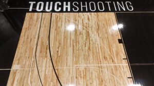 Overhead view of a basketball court with wood floor and white boundary lines; large white letters spelling 'TOUCH SHOOTING' along a black border at the top/border.