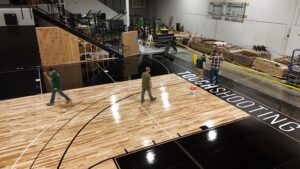 Indoor gym construction site with a partially finished wooden basketball court and workers moving equipment in the background.