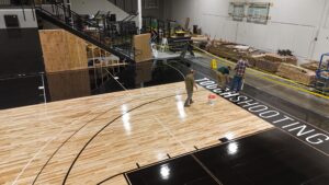 Construction workers install a new hardwood gym floor, with yellow caution tape and building materials nearby in a large indoor space.