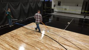 Two workers mop a basketball court; one in a green shirt with blue gloves, the other in a plaid shirt pushing a mop across the wooden floor.
