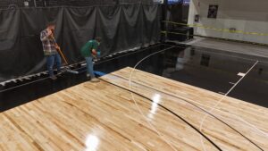 Two workers on an indoor basketball court finish a glossy black epoxy floor, one using a floor squeegee and the other laying tape along the edge.