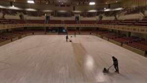 Empty theater auditorium with red seats; workers polish the wooden stage floor using floor polishing machines.