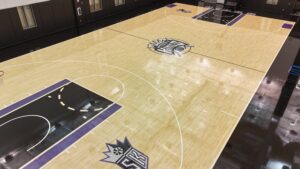 Empty indoor basketball court with a light wood floor, purple and black accents, and a large team logo at midcourt.