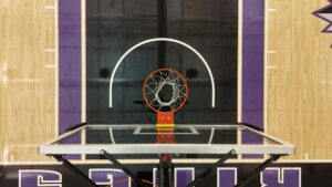 Basketball hoop from below in a gym, orange rim and white net against a dark backboard with purple wall padding surroundings.
