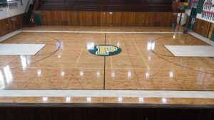 High-angle view of a school gymnasium basketball court with a green circle center logo featuring a yellow 'P', wood floor, and bleachers in the background, plus a person kneeling near the right wall.