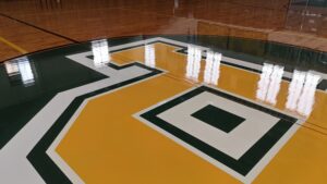 Close-up of a polished gym floor featuring a large green, white, and yellow logo or letter at center court.