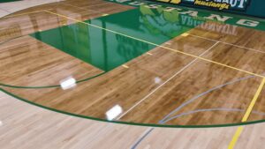 Indoor basketball court with a polished wood floor, green center area, and yellow/white boundary lines.