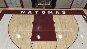 Overhead view of a high school basketball court with 'NATOMAS' painted in bold white letters across a maroon center stripe.