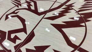 Circular maroon emblem with angular zigzag patterns painted on a light hardwood gym floor (basketball court).
