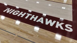 Indoor basketball court floor with a maroon stripe bearing large white letters spelling 'NIGHTHAWKS'.
