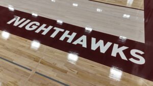 Indoor basketball court floor with a maroon central stripe displaying the word 'NIGHTHAWKS' in large white letters