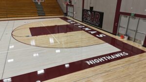 Empty school gym with polished hardwood floor, maroon sidelines reading 'NIGHTHAWKS' beside the court and bleachers to the left