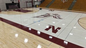 Empty school gym with polished wood floor, a lone person kneeling near the center court, Natomas High School logo on the wall, and empty wooden bleachers to the right.