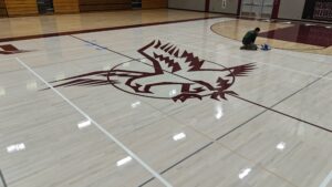 Maintenance worker kneels on a polished gym floor, using a buffer near a large maroon logo in the center court.