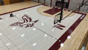 Worker kneels on a polished high school basketball court, painting or taping lines while a large school logo is visible on the floor.