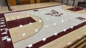 Indoor high school basketball court with maroon and beige wood flooring, center logo of a stylized bird, and bleachers along the far wall.