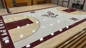 Empty Natomas High School gymnasium with a polished wooden basketball court, maroon borders, and a central mascot logo.