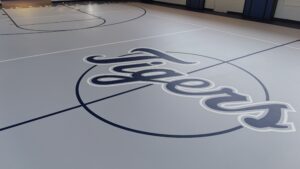 Indoor gym floor featuring a large dark blue cursive logo painted on light gray surface, with basketball court lines in the foreground and walls in the background.