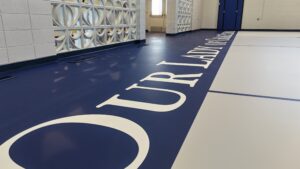 Wide view of an indoor gym floor with a dark blue strip bearing large white letters, next to light gray flooring and a decorative wall.