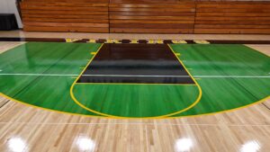 Indoor gymnasium basketball court with green flooring, yellow boundary lines, and 'VIKINGS' along the sideline near wooden bleachers.