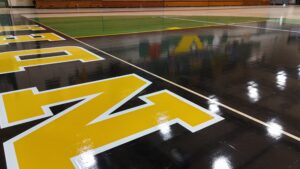 Polished gymnasium floor with large yellow block lettering and white boundary lines, reflections visible on the surface.