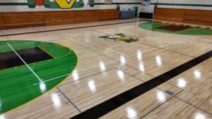 Empty school gym with polished wood floor, green/yellow court markings, and a central mascot logo surrounded by bleachers on both sides.