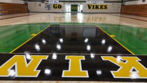 Indoor school gym with a polished floor, green and black court in the foreground, and large yellow letters spelling 'VIKES' along the baseline; bleachers along the sides and banners on the back wall.