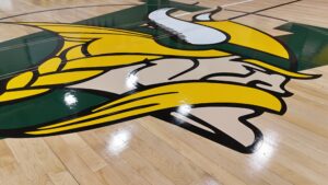 Large mascot graphic painted on a polished basketball court floor, featuring yellow, white, and green colors.